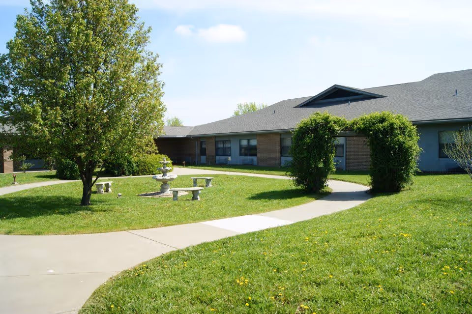 Outdoor view of a senior living facility with a curved concrete pathway, green grass, a tree, a small fountain surrounded by stone benches, and a building with multiple windows in the background. There is also a green archway made of bushes along the pathway.