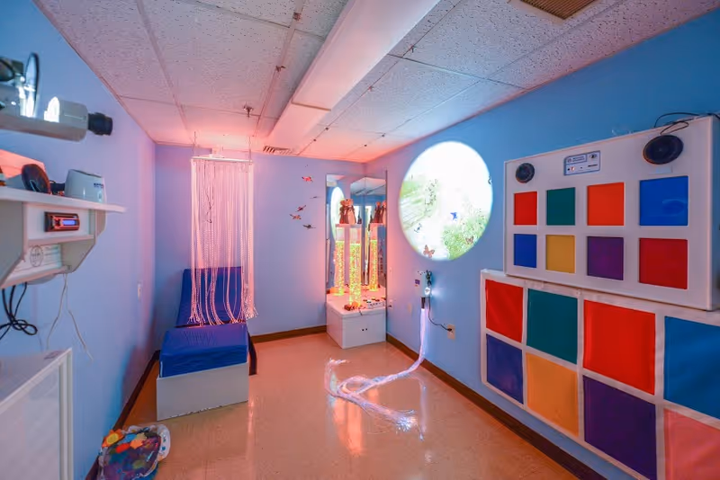 Blue sensory therapy room in St. Mary's with seating, colorful panels, circular window.