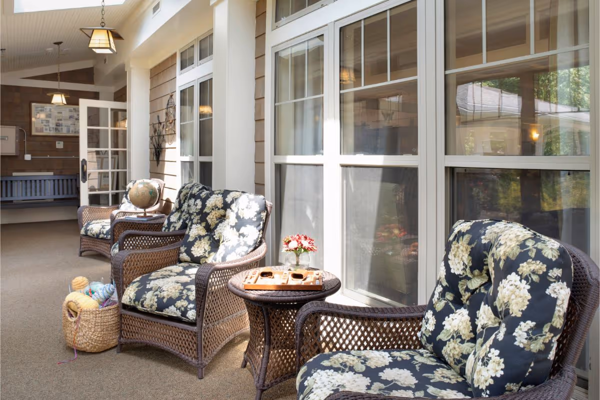 Sunlit enclosed patio-style sitting area with wicker chairs upholstered in floral cushions, side tables, and a basket of yarn.