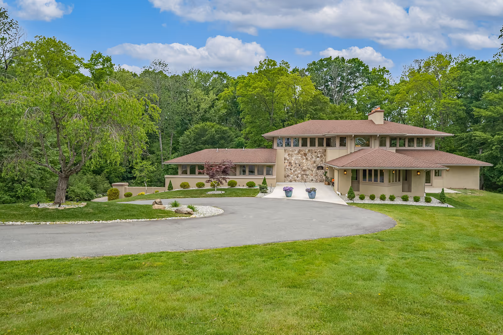 A large, modern two-story house with a stone facade and brown roof surrounded by green grass and trees under a partly cloudy sky.