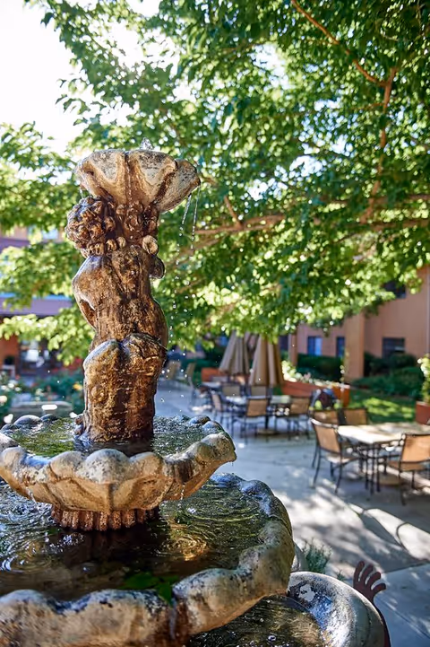 A close-up view of a decorative stone water fountain with water gently flowing from the top tier. In the background, there is an outdoor patio area with tables, chairs, and umbrellas surrounded by greenery and trees providing shade.