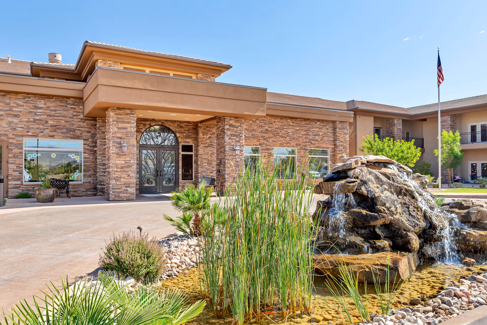 Exterior view of The Retreat at Sunriver building entrance with stone facade, decorative wrought iron double doors, a small pond with a rock waterfall feature, and landscaping including tall grasses and palm plants under a clear blue sky.