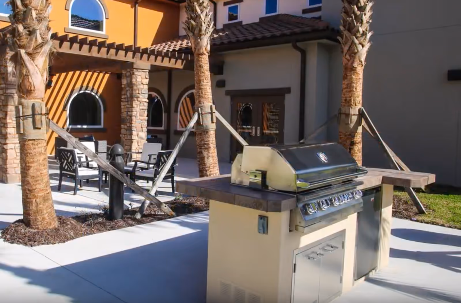 Patio courtyard at a senior living facility featuring a stainless-steel outdoor grill, seating area, palm trees, and an arched building facade.