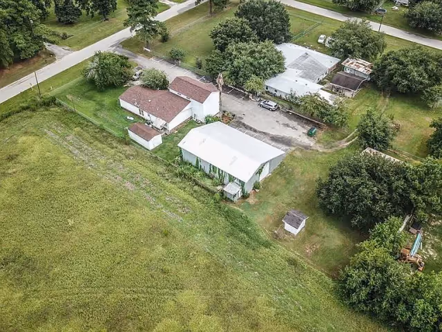 Aerial view of a small facility with several buildings surrounded by green grass and trees. There is a paved road nearby and a few parked vehicles. The area appears rural with open fields and scattered trees.