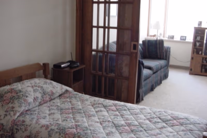 A bedroom with a wooden bed frame and a floral patterned quilt. Next to the bed is a small wooden nightstand with a phone on it. A wooden sliding door with glass panels separates the bedroom from a living area, which contains a striped couch, a window with a lamp on the sill, and a wooden cabinet with shelves.