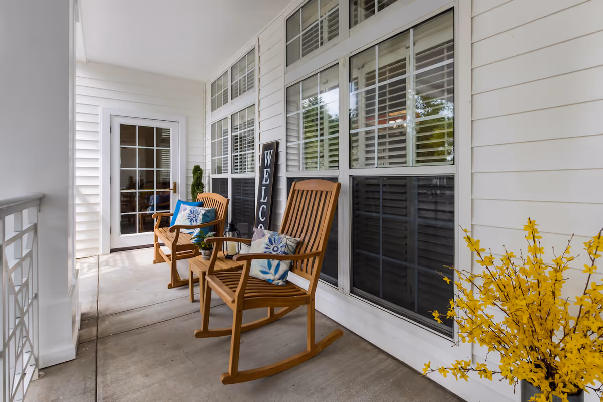 A covered porch area with two wooden rocking chairs, each with a decorative cushion, a small wooden table between them holding a small plant and a lantern, a tall welcome sign leaning against the wall, white siding and large windows with white shutters, and a pot of yellow flowers on the right side.