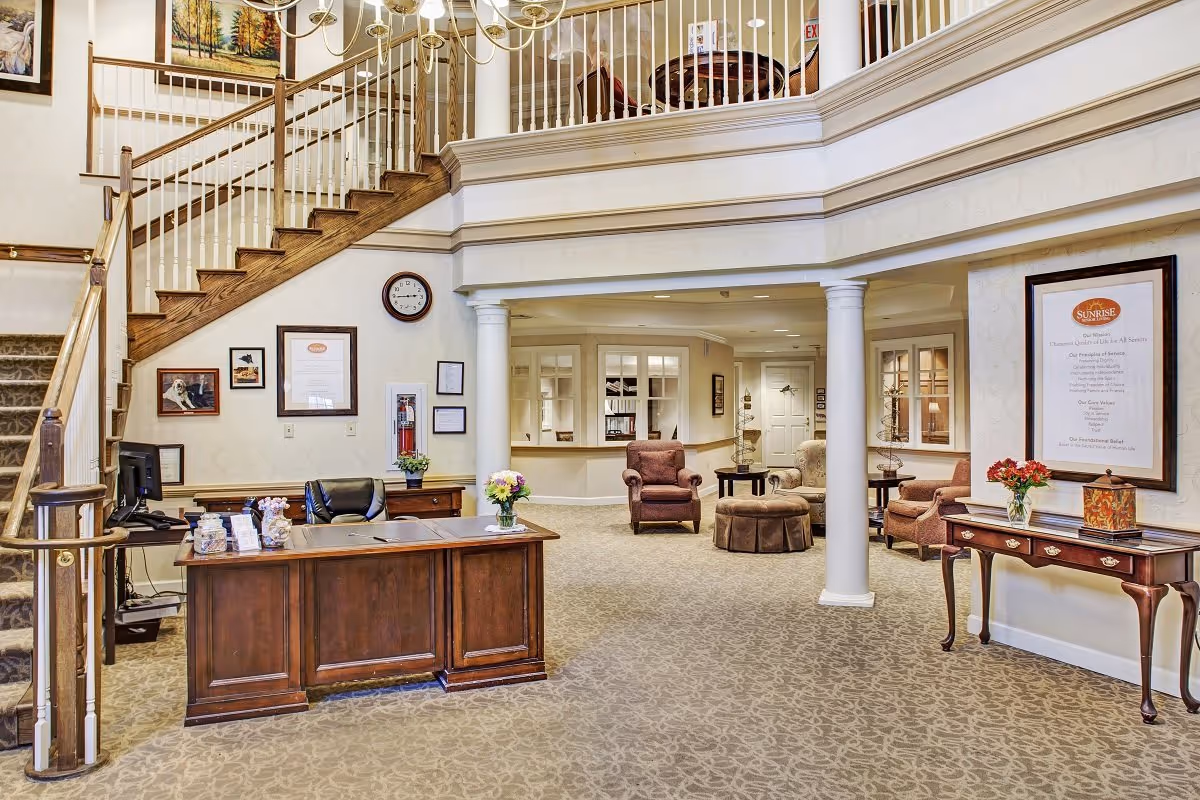 Interior view of a senior living facility lobby with a wooden reception desk, a staircase with wooden handrails and white balusters, a clock on the wall, framed pictures and certificates, and a seating area with armchairs and a round ottoman. The space has beige patterned carpet and cream-colored walls with decorative molding.