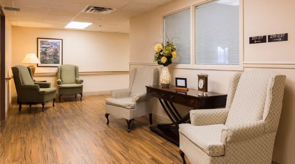 A cozy hallway seating area in Sanders Glen Assisted Living Center featuring four upholstered armchairs, two green and two beige, arranged around a wooden console table with a vase of flowers, a clock, and a framed picture. The floor is wood, and there are directional signs on the wall indicating room numbers.