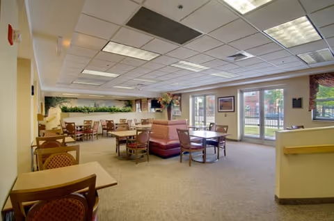 Spacious senior living dining room with multiple tables and chairs, carpeted floor, and large windows/doors letting in natural light.