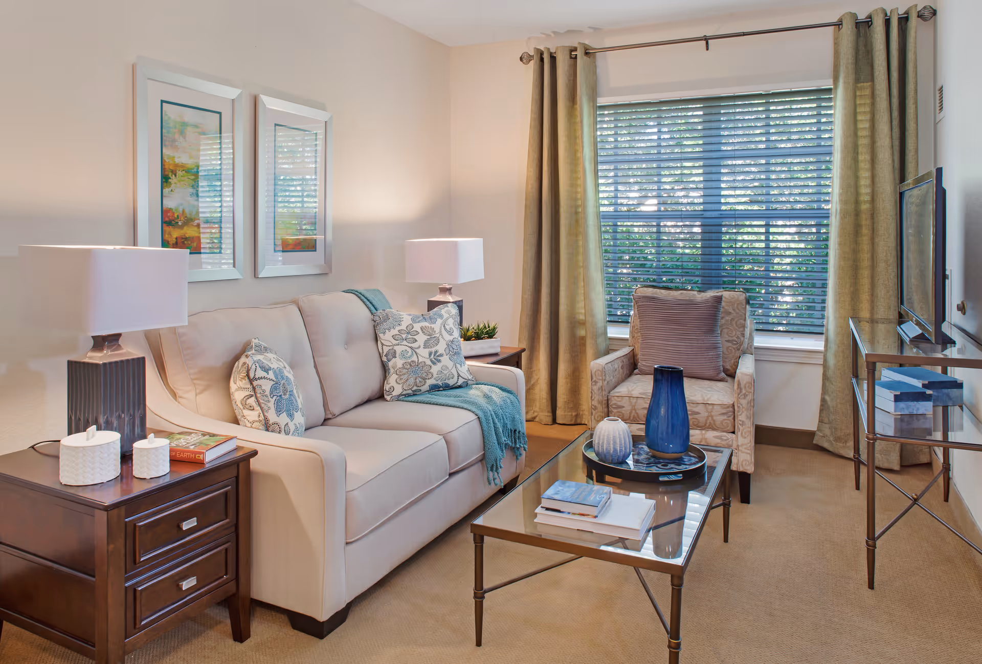 A cozy living room with a beige sofa adorned with patterned cushions and a teal throw blanket. Next to the sofa is a dark wooden side table with two white decorative containers and a book. A glass coffee table holds a blue vase, a white vase, and some books. An armchair with a patterned fabric and a purple cushion sits near a window with beige curtains and horizontal blinds. A flat-screen TV is mounted on a metal and glass stand to the right.