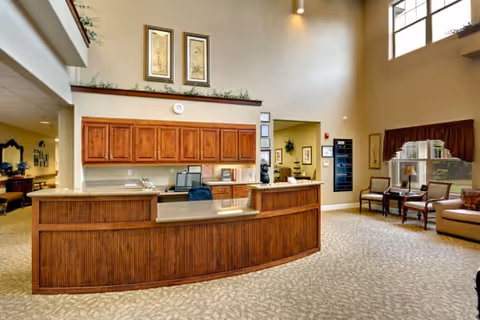 Interior view of a senior living facility reception area with a curved wooden front desk, office equipment, and seating area with chairs and a sofa near large windows. The walls are decorated with framed artwork and plants.