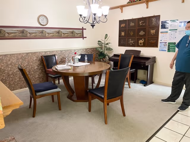 A small dining area with a round wooden table surrounded by four chairs. On the table are some papers, a flower in a vase, and hand sanitizer bottles. The room has a wall clock, decorative wall art, a potted plant, and a piano against the wall. A person wearing a mask and teal shirt stands near the right side of the image.