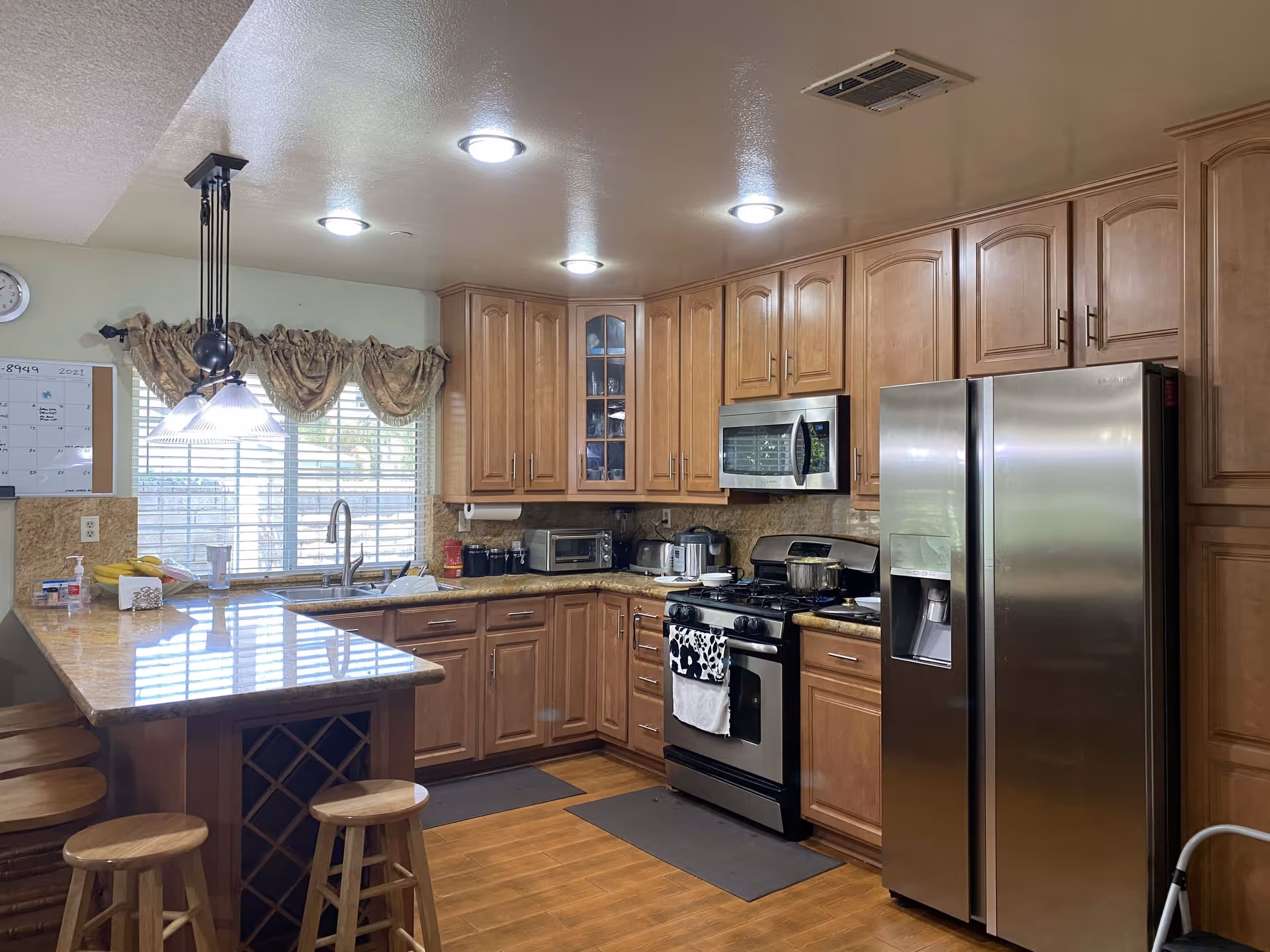 A well-lit kitchen with wooden cabinets, a stainless steel refrigerator, microwave, and stove. There is a granite countertop with four wooden stools, a window with blinds and a decorative curtain, and various kitchen appliances and items on the counters.
