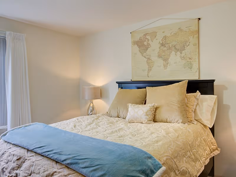 Neatly made bed in a softly lit bedroom with beige bedding, a blue throw, pillows, a bedside lamp, and a world map hanging above the headboard.