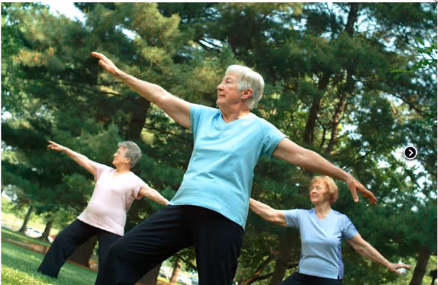 Three elderly women practicing tai chi or a similar stretching exercise outdoors in a park-like setting with green trees in the background.
