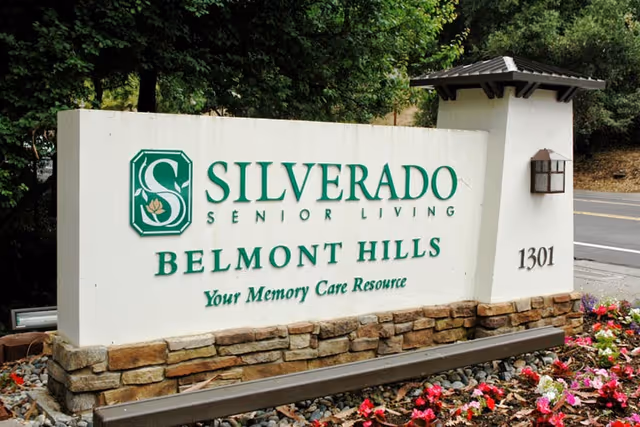Outdoor sign for Silverado Senior Living Belmont Hills, Your Memory Care Resource, with the address number 1301. The sign is white with green text and a stone base, surrounded by flowers and greenery.