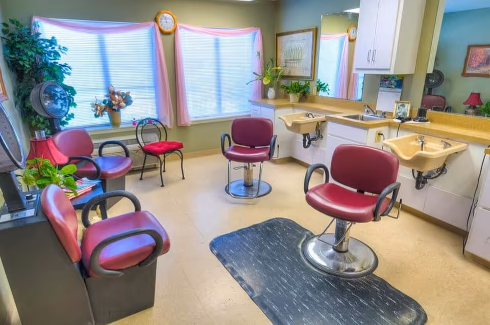 Small bright salon room with multiple maroon styling chairs, shampoo sinks, mirrors, and potted plants.