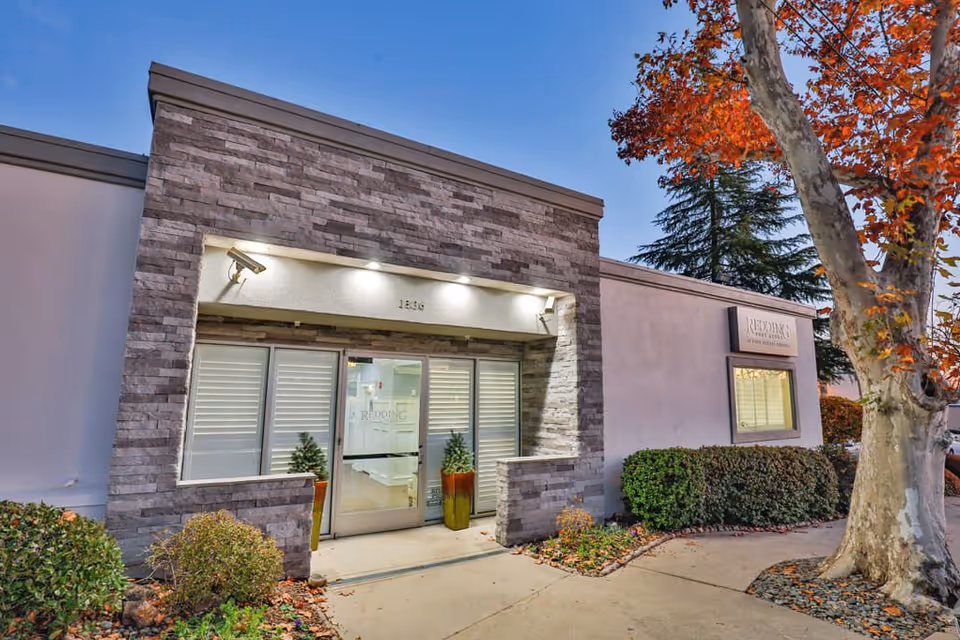 Exterior entrance of Redding Post Acute facility with stone facade, glass double doors, potted plants on either side of the entrance, and surrounding landscaping including bushes and a tree with autumn leaves.