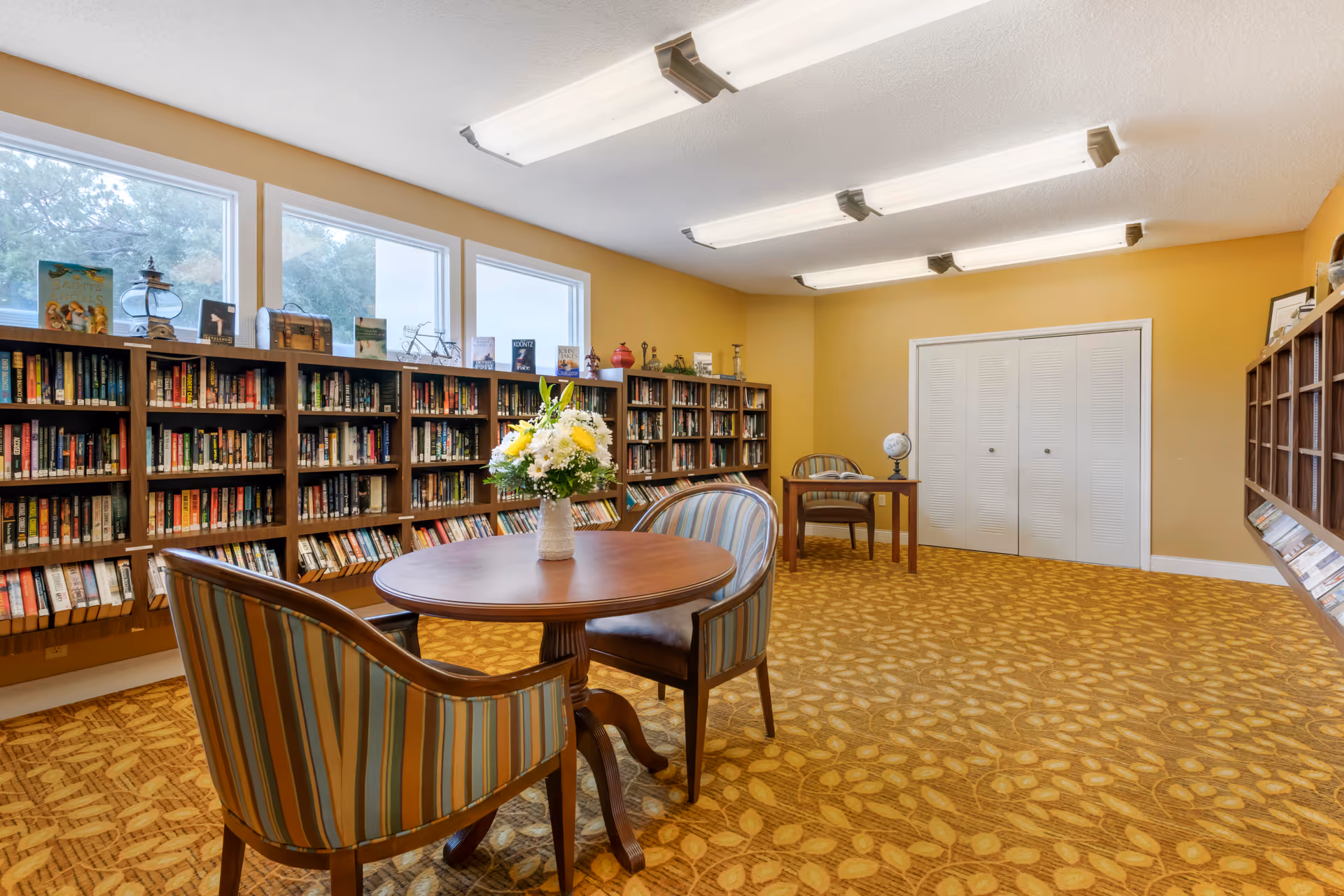 A bright library/reading room with round tables, striped chairs, bookshelves lining the walls, and a vase of flowers on the table.