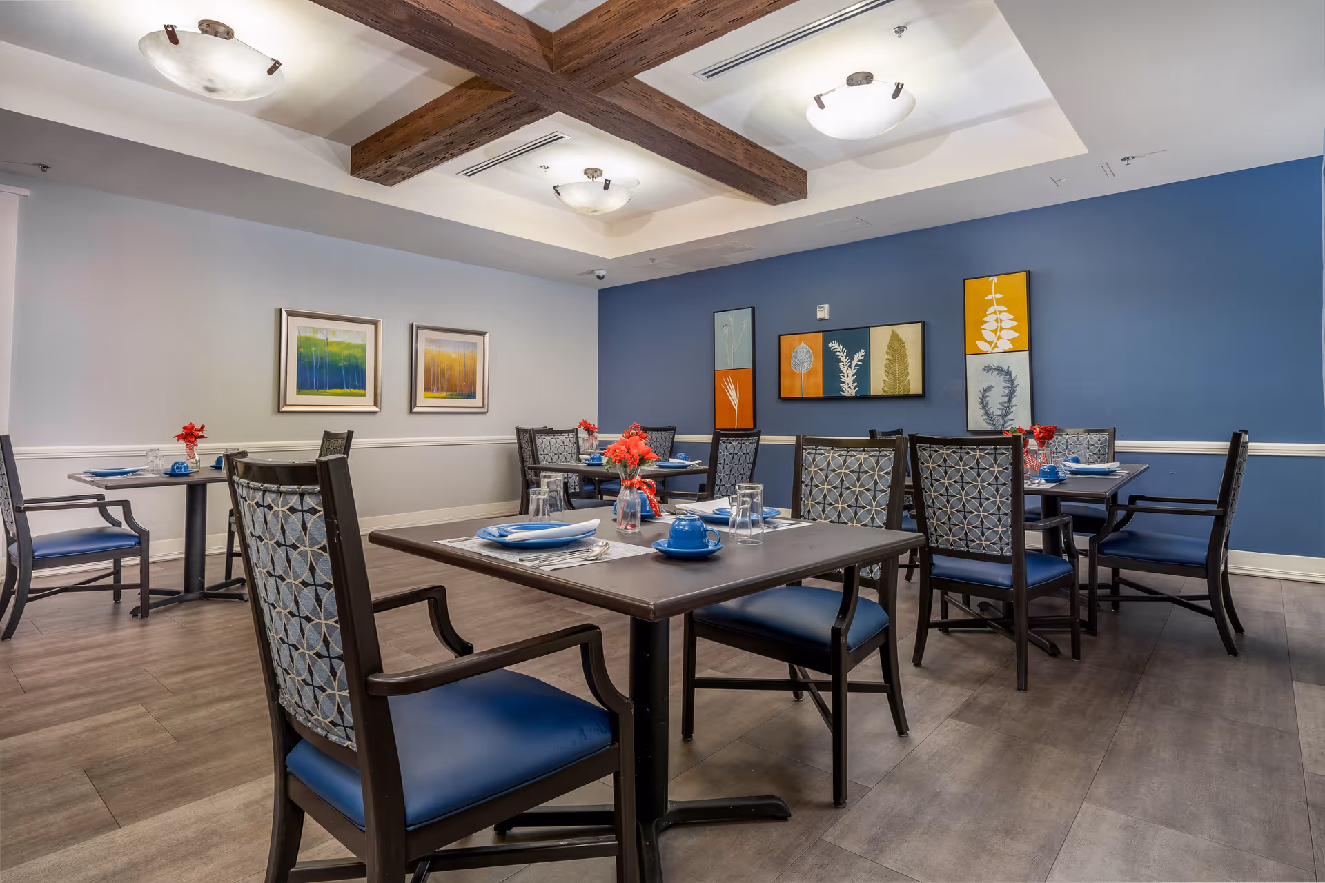 Dining room with several set tables and blue-upholstered chairs, decorative artwork on blue walls, and exposed wooden ceiling beams.