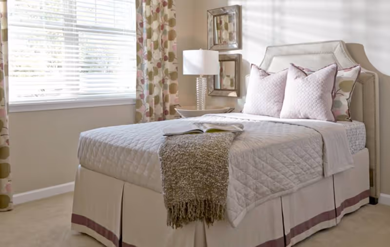 A neatly made bed with a quilted beige bedspread, three decorative pillows, and a textured throw blanket at the foot. The bed is positioned next to a window with floral patterned curtains. A bedside table with a lamp and two square mirrors on the wall are also visible.