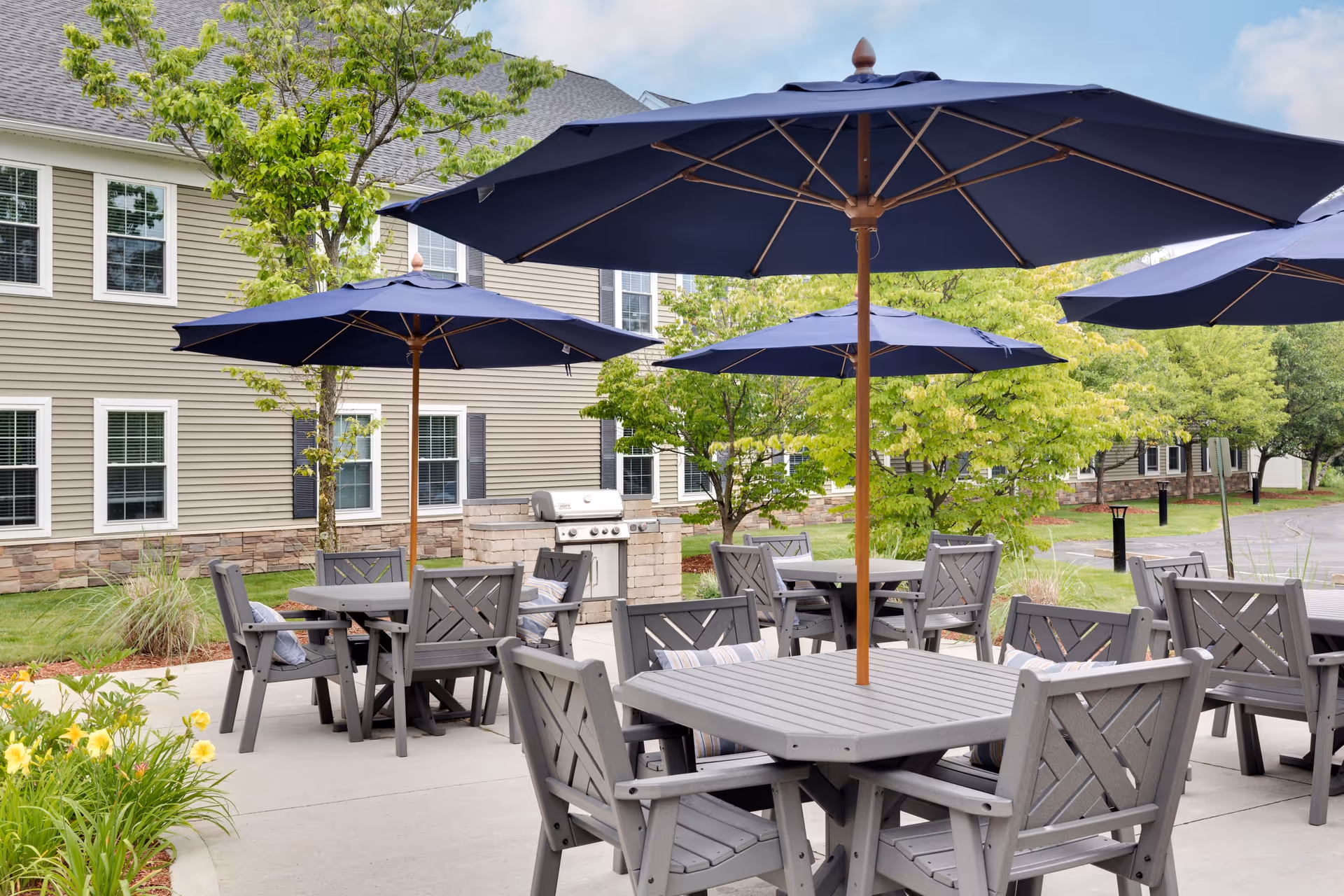 Outdoor patio area with multiple gray tables and chairs, each table shaded by a large blue umbrella. There are green trees and plants surrounding the patio, and a building with beige siding and white-trimmed windows in the background. A stainless steel grill is also visible near the building.