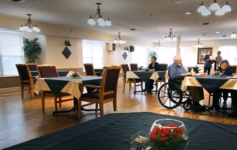 Dining room in a senior living facility with tables set with tablecloths and several elderly residents seated, including a man in a wheelchair.