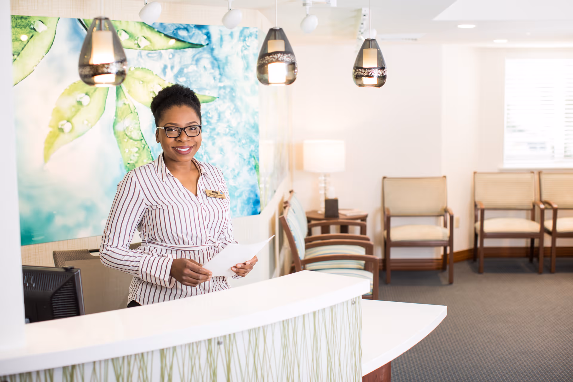 A smiling woman wearing glasses and a striped shirt stands behind a white reception desk holding papers. Behind her is a colorful wall mural with green leaves and blue background. The waiting area with several chairs and a side table with a lamp is visible in the background.