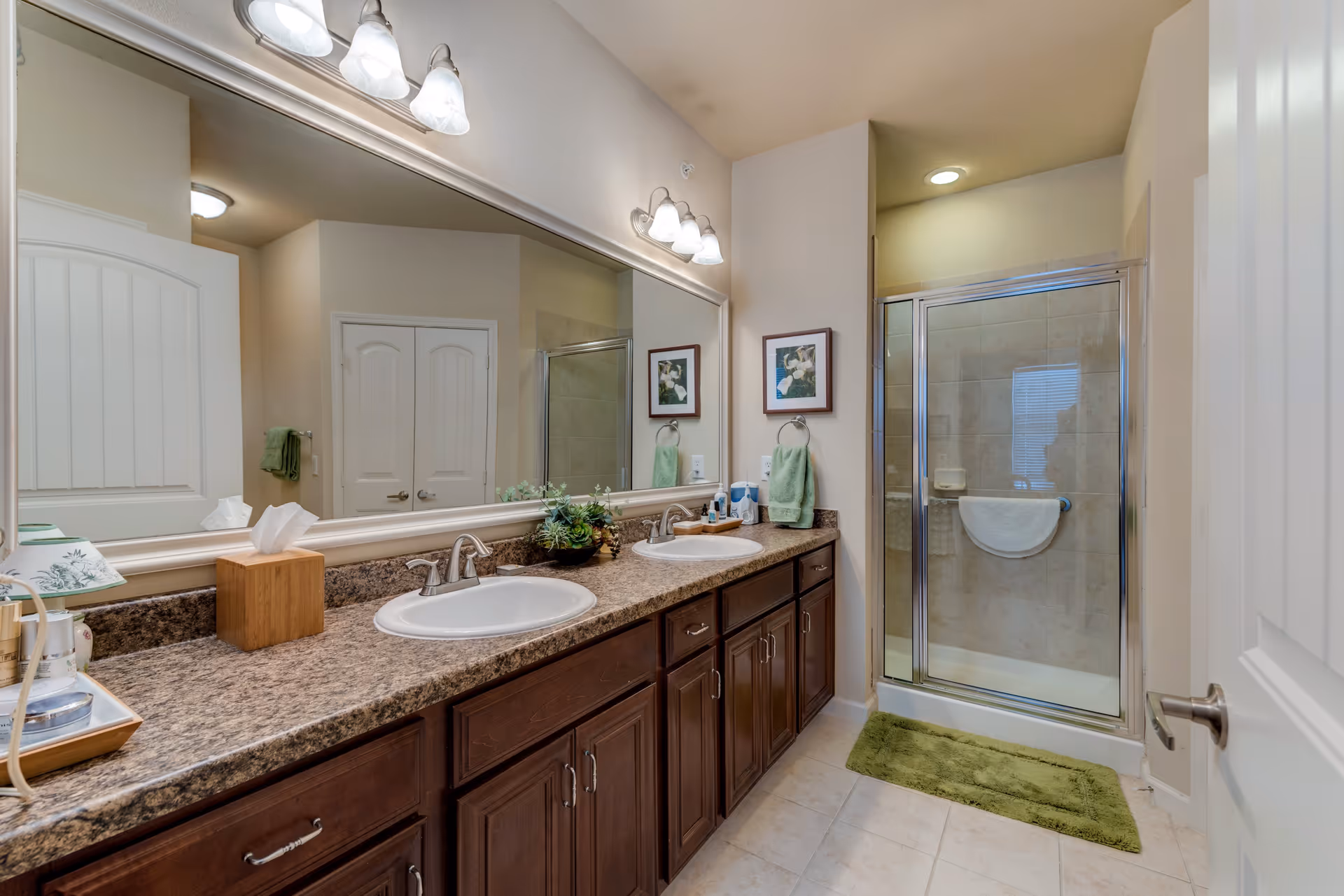 A clean and well-lit bathroom featuring a double sink vanity with dark wood cabinets and a granite countertop. Above the sinks is a large mirror with three-light fixtures. To the right is a glass-enclosed shower with beige tile walls and a green bath mat on the floor. The walls are painted a light beige color and there are framed pictures and green towels hanging on the wall.