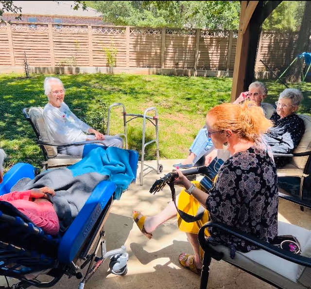 A group of elderly people sitting outside in a shaded patio area with a wooden fence and green grass in the background. One woman with red hair is playing a guitar while others listen and relax in chairs and a wheelchair.