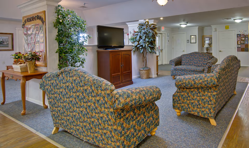 A senior living facility common area with three floral-patterned upholstered sofas arranged around a wooden TV stand holding a flat-screen television. The room has carpeted flooring with a wooden floor border, potted plants, a wooden side table with flower arrangements, and a bulletin board on the wall.
