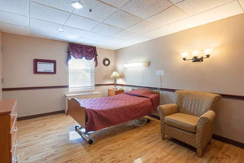 A simple, clean bedroom in a healthcare facility featuring a single bed with a maroon bedspread, a wooden nightstand with a lamp, a comfortable patterned armchair, a window with blinds and a maroon valance, and light wood flooring.