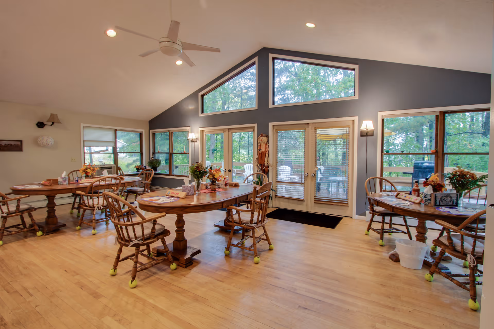 A bright dining room with wooden tables and chairs, each chair fitted with tennis balls on the legs. The room has large windows and glass doors that provide a view of green trees outside. The walls are painted gray and white, and the ceiling is vaulted with a white ceiling fan and recessed lights. The tables are decorated with flowers and placemats, and there is a scarecrow decoration near the glass doors.