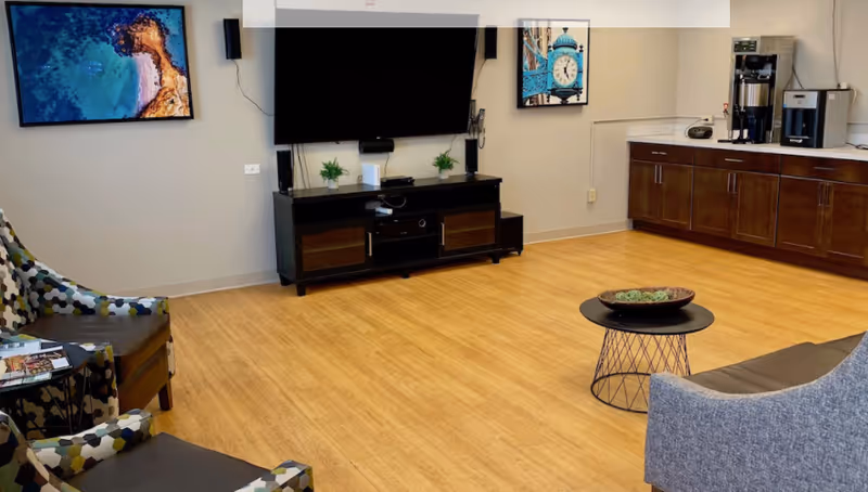 A cozy living room area with a wall-mounted flat screen TV above a black entertainment console, two patterned armchairs, a gray sofa, a small round black coffee table with decorative items, wooden flooring, and a countertop with a coffee machine and cabinets. Two framed pictures hang on the walls.
