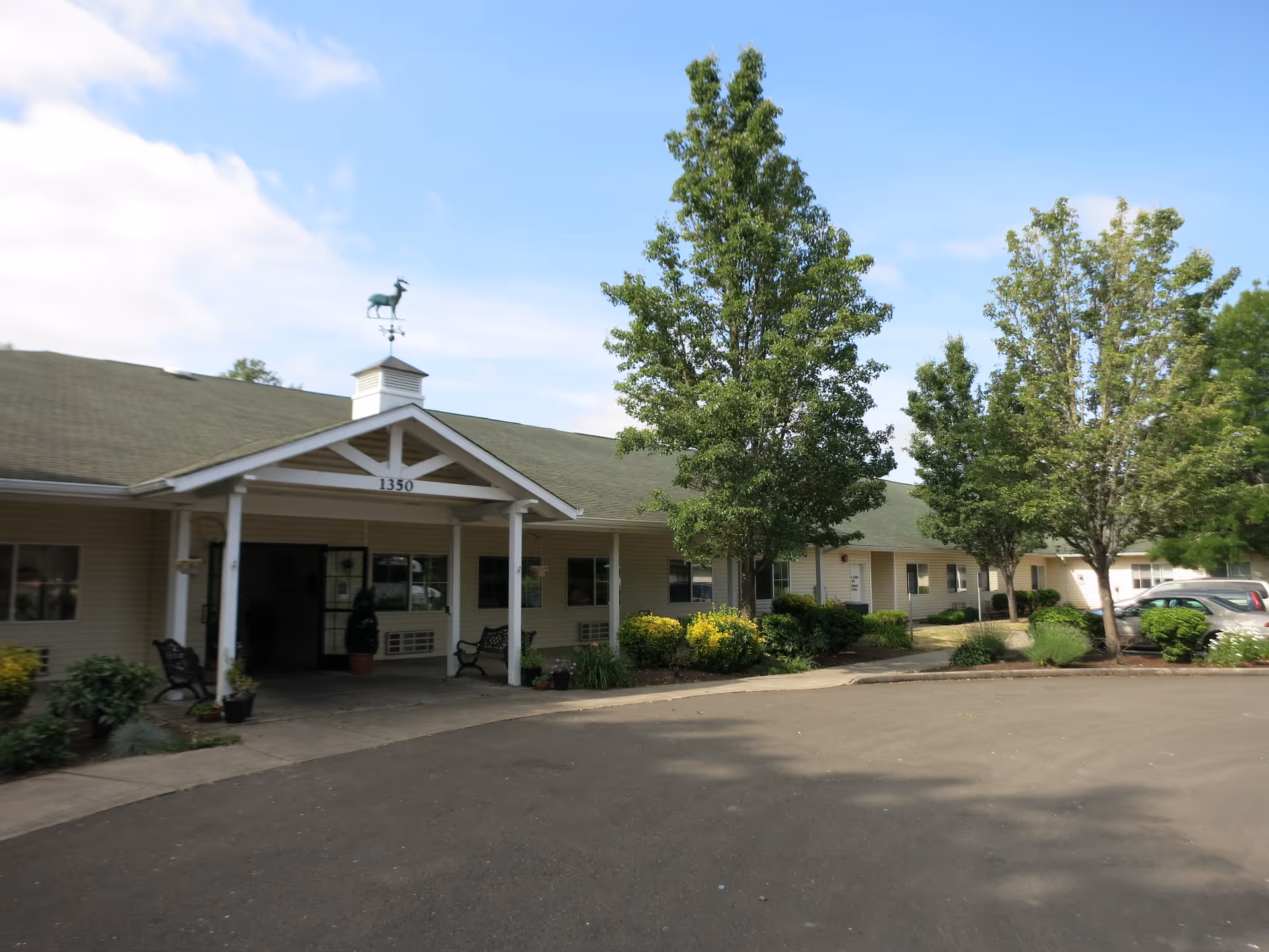 Exterior view of a single-story senior living facility building with a covered entrance, benches, and a weather vane on the roof. The building is surrounded by trees, shrubs, and a paved driveway under a partly cloudy sky.