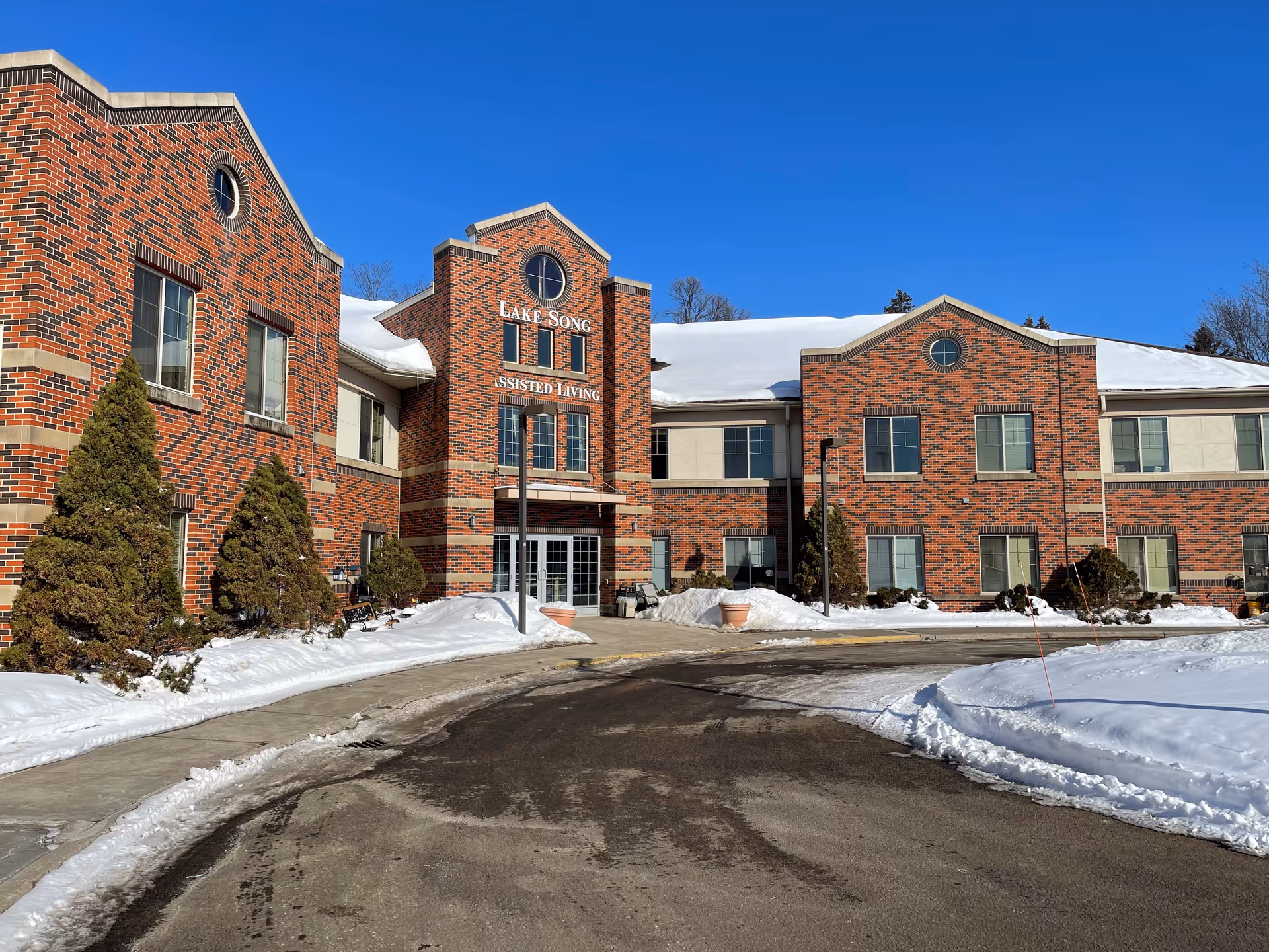 Exterior view of a two-story brick building with snow on the ground and roof under a clear blue sky. The building has multiple windows and a central entrance with the sign 'Lake Song Assisted Living' above the door. There are small evergreen trees and shrubs along the sidewalk leading to the entrance.