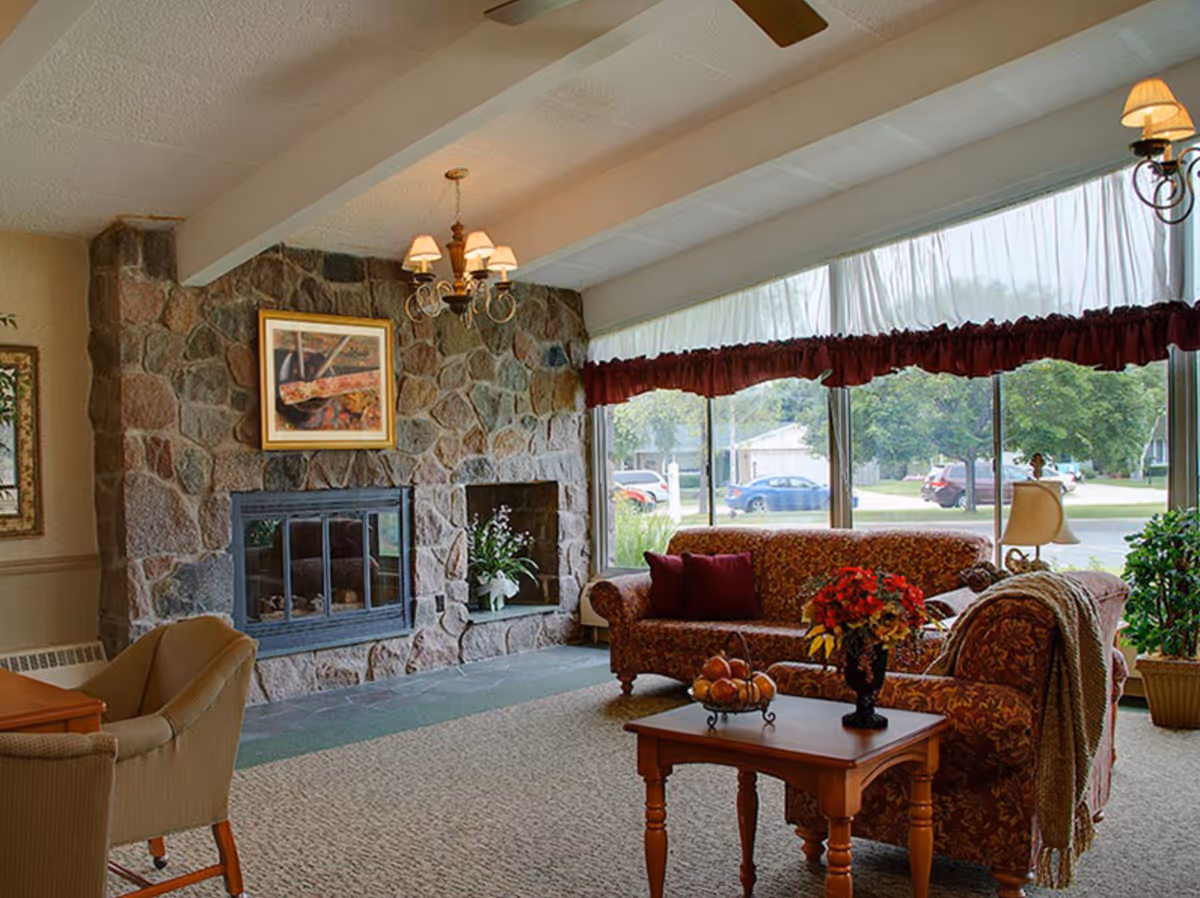 Cozy common living room with a stone fireplace, patterned sofas, a coffee table with flowers, and large windows overlooking the street.
