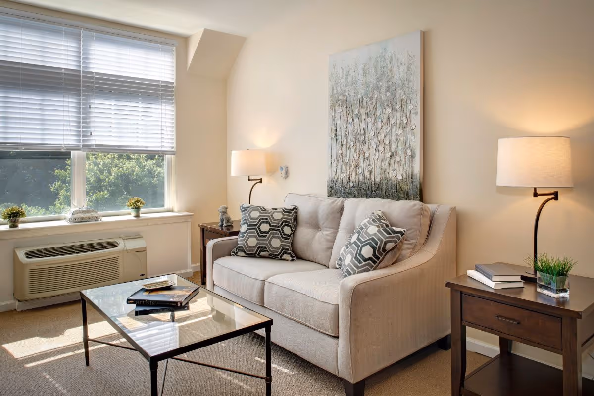 A cozy living room with a beige sofa adorned with two patterned cushions, a glass coffee table with books on it, two wooden side tables each with a lamp and decorative items, and a large window with white blinds letting in natural light. A large abstract painting hangs on the wall above the sofa.