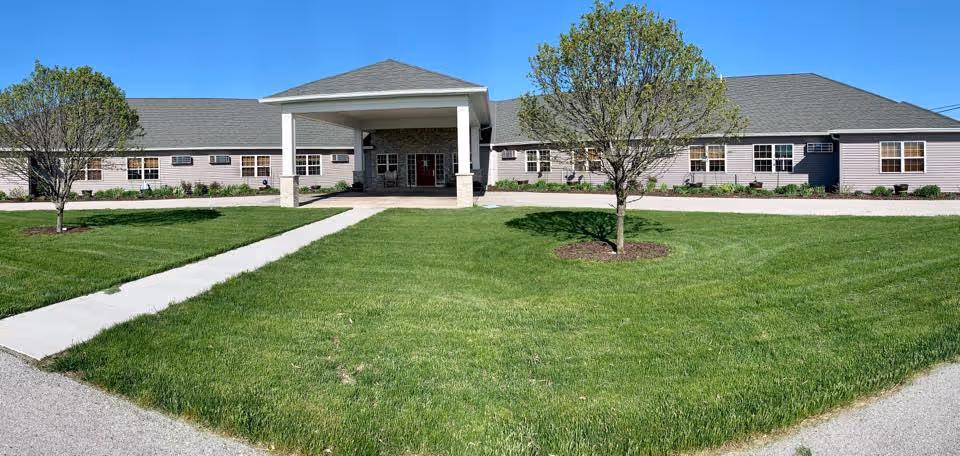 Front exterior view of a single-story assisted living facility building with a covered entrance, surrounded by green grass and a few trees under a clear blue sky.