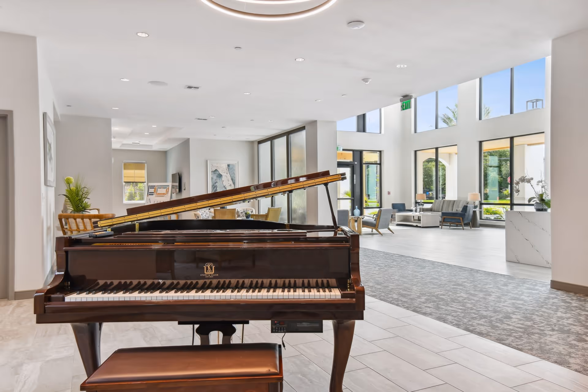Interior view of a senior living facility lobby featuring a grand piano in the foreground, with seating areas and large windows allowing natural light to fill the space.