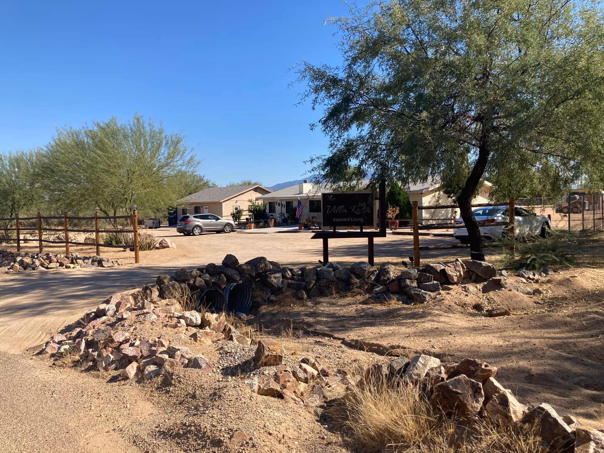 Exterior view of The Villa Rosa Assisted Living facility showing a single-story building with parked cars, a wooden fence, desert landscaping with rocks and trees, and a clear blue sky.