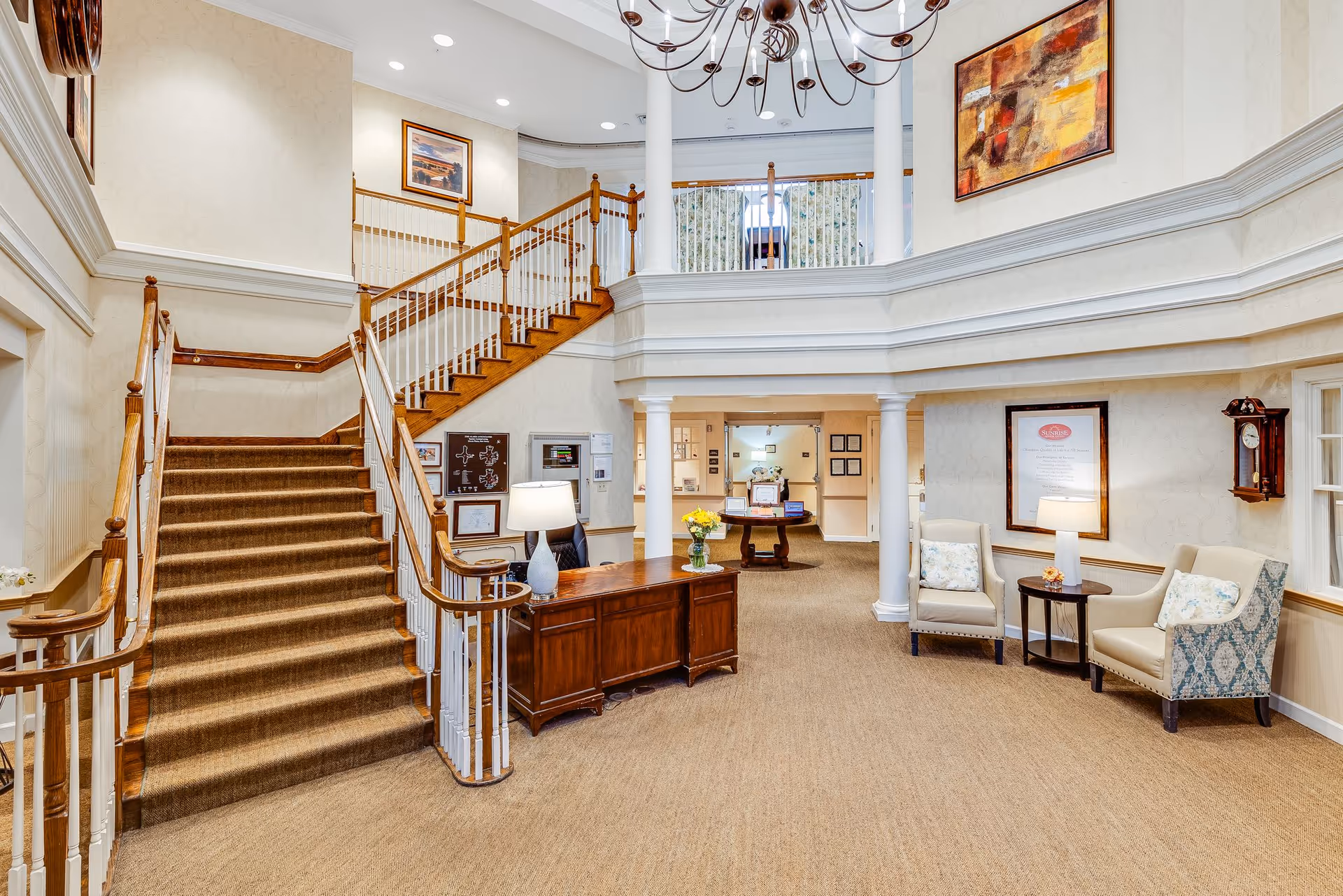 Interior view of a senior living facility lobby with a carpeted staircase leading to an upper level. There is a wooden reception desk with a lamp and flowers, two upholstered chairs with a small table between them, and framed artwork on the walls. The space is well-lit with ceiling lights and a chandelier.