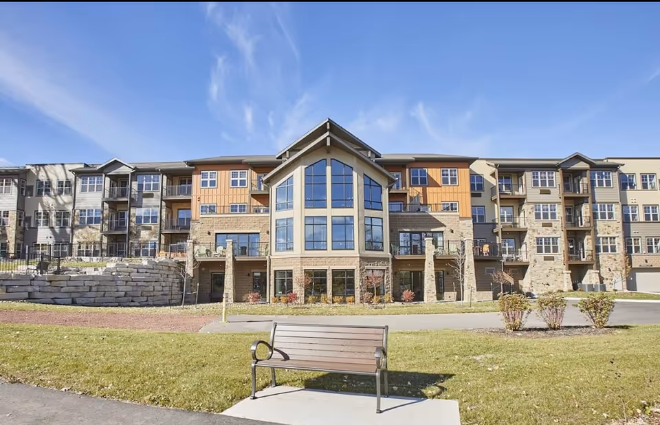 Exterior view of a modern multi-story senior living facility with large windows and balconies. In the foreground, there is a wooden bench on a concrete pad surrounded by grass and small landscaped bushes under a clear blue sky.