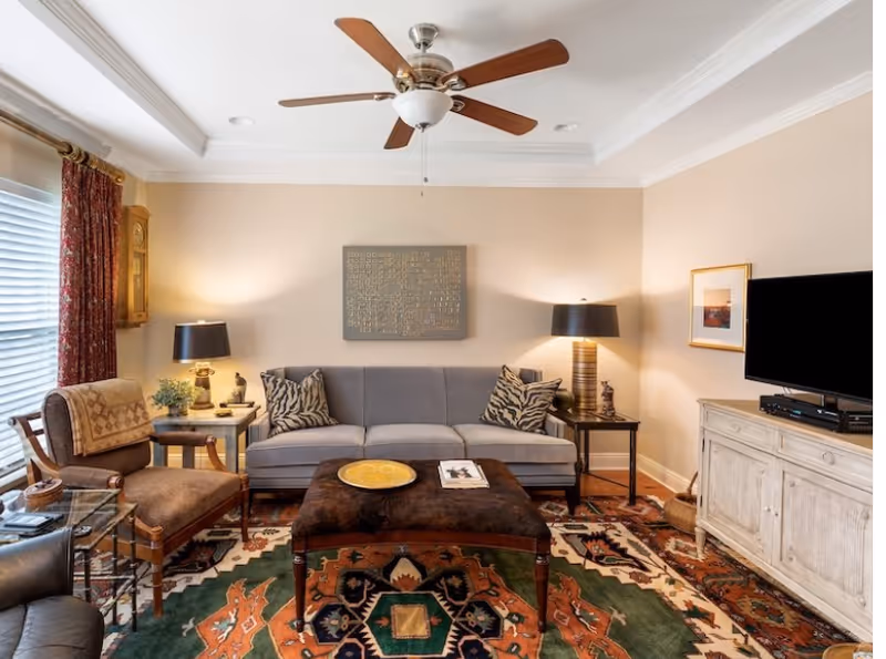 A cozy living room with a gray sofa adorned with patterned pillows, two side tables each with a lamp, a large patterned rug, a wooden armchair with a cushion, a ceiling fan with wooden blades, a flat-screen TV on a white wooden cabinet, and a window with red patterned curtains.