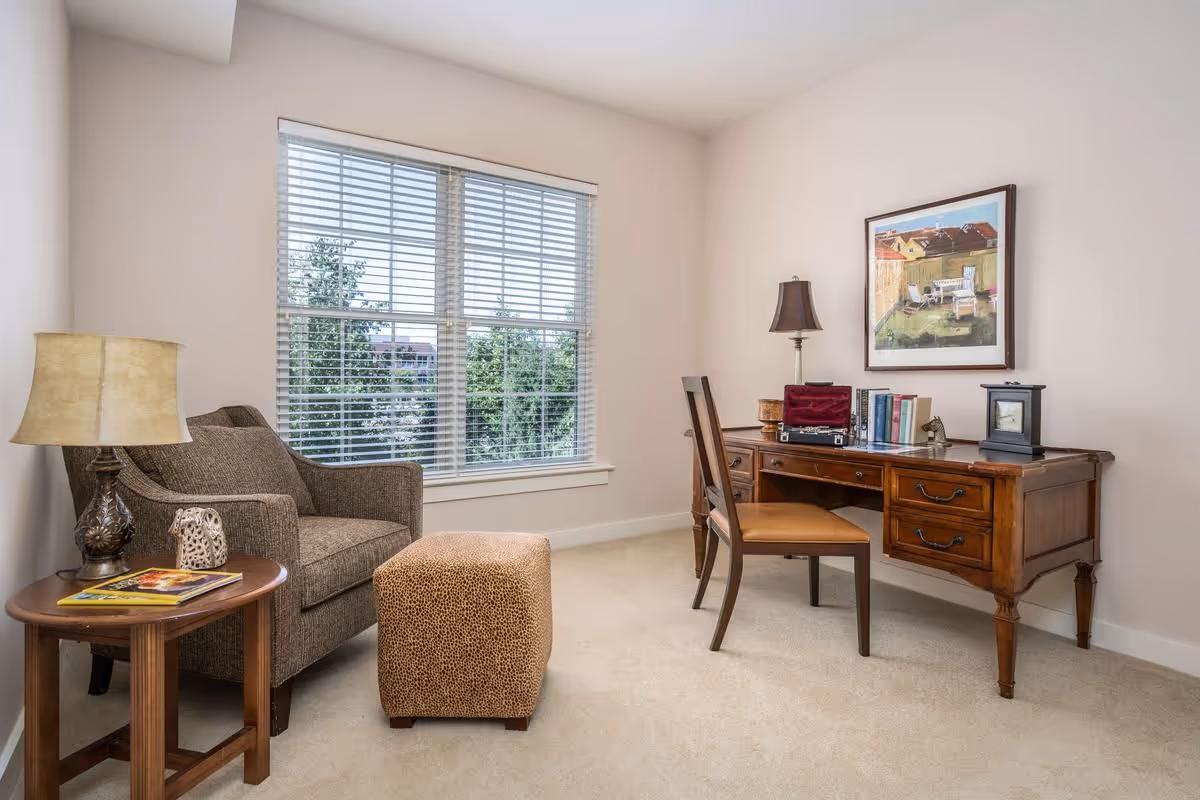 A cozy room with a large window covered by white blinds, a brown upholstered armchair next to a wooden side table with a lamp and decorative items, a leopard print ottoman, and a wooden desk with a chair, books, a lamp, and a framed painting on the wall above it.