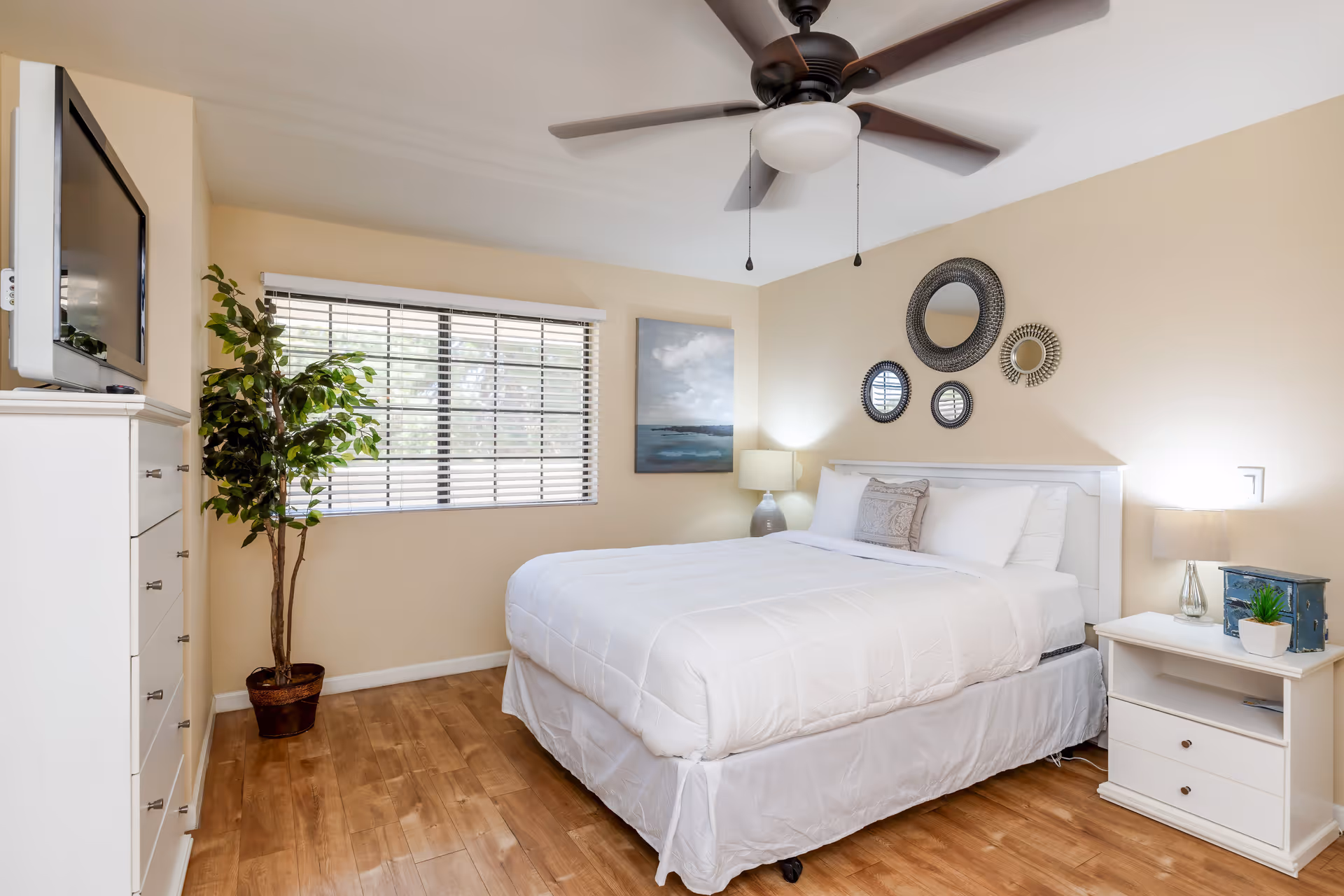 A bright and clean bedroom with a white bed, white bedding, and a decorative pillow. The room features a ceiling fan with lights, a window with blinds, a potted plant, a white dresser with a TV on top, and a white nightstand with a lamp and small decorative items. Circular mirrors and a painting hang on the beige walls.