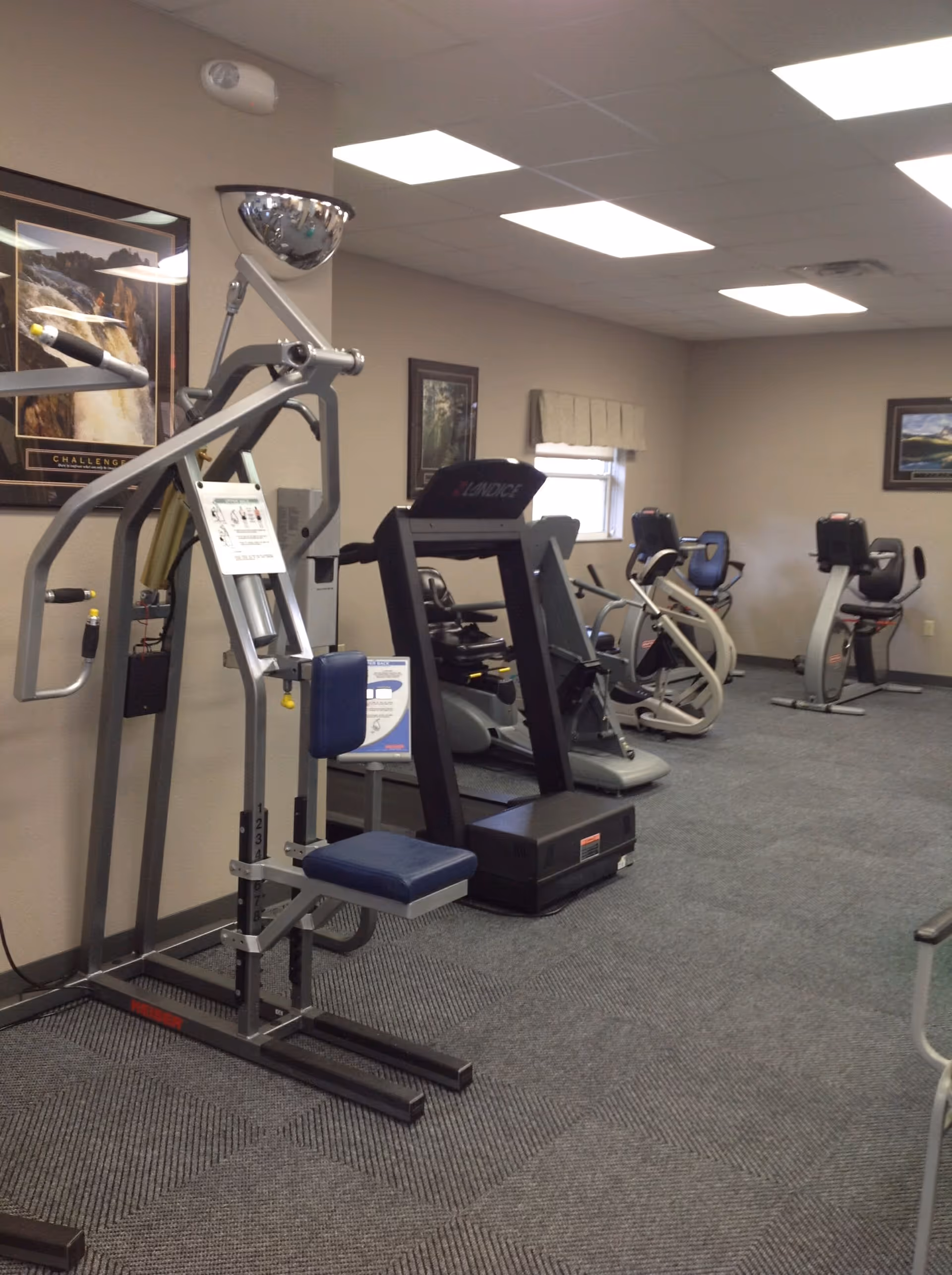 Indoor fitness room with various exercise equipment including a weight machine, treadmill, and stationary bikes. The room has carpeted flooring, beige walls, framed pictures on the walls, and a window with a valance.