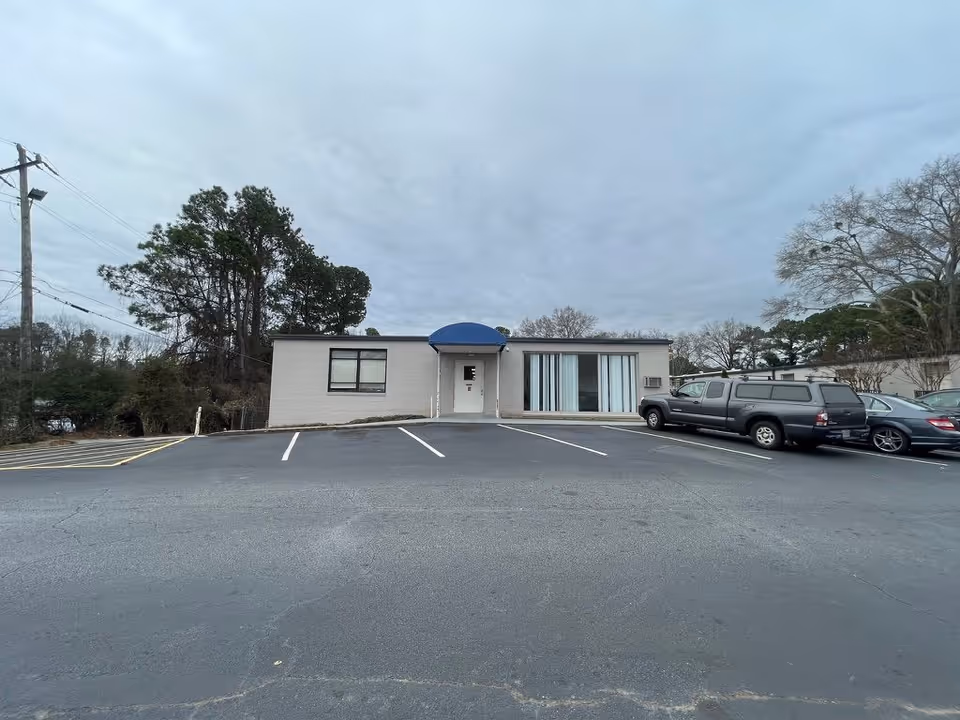 Single-story brick building with a blue awning and sliding glass windows set behind a mostly empty parking lot on an overcast day.