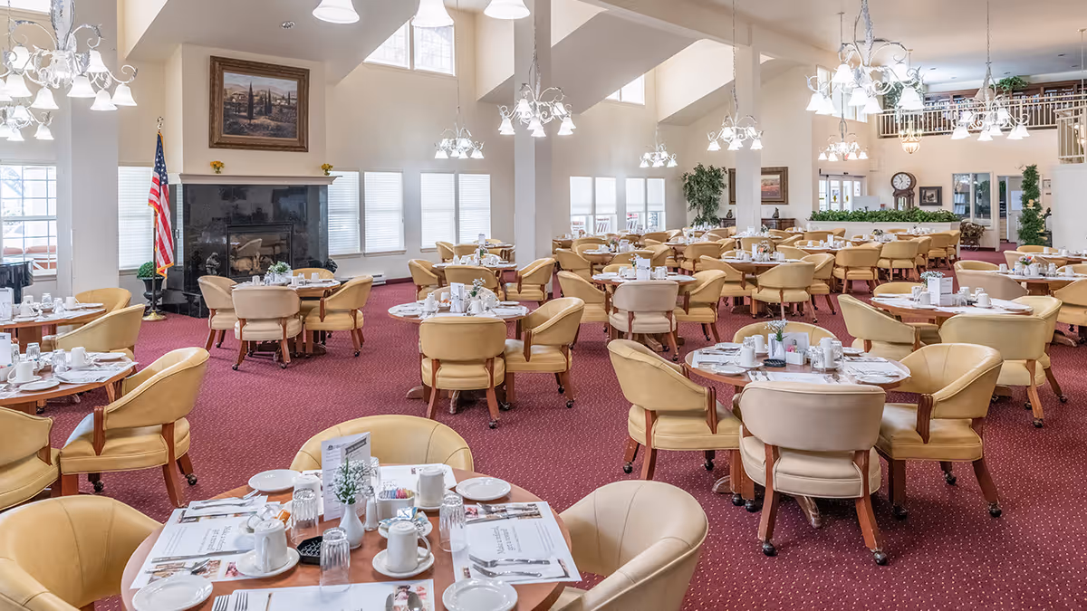 Spacious dining room with multiple round tables set with white cups, plates, and utensils. The room has beige cushioned chairs, red carpet flooring, large windows with blinds, chandeliers hanging from a high ceiling, and a fireplace with an American flag beside it.