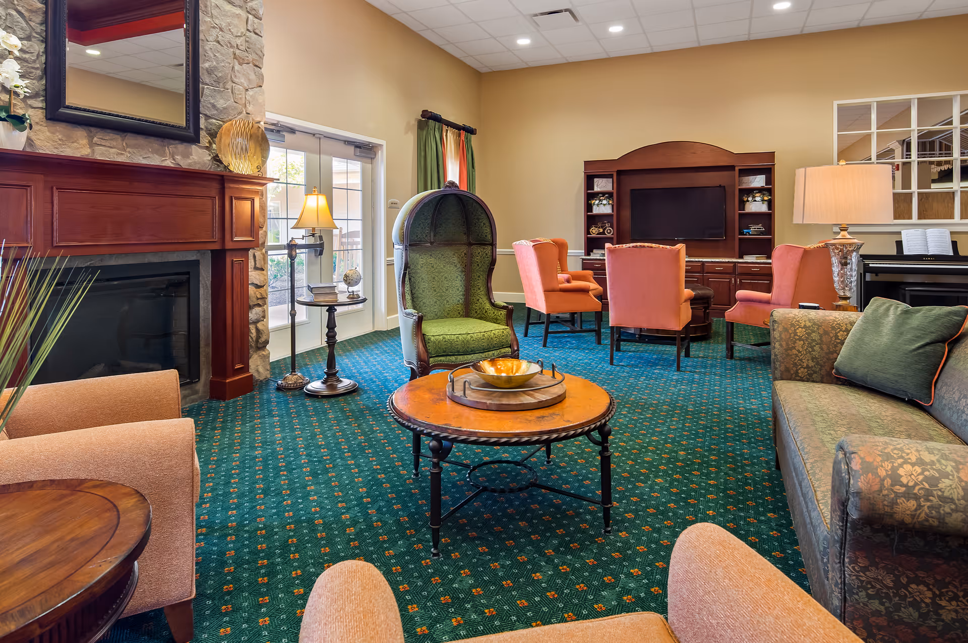 A cozy living room area in a senior living facility featuring a stone fireplace with a wooden mantle, a large mirror above it, and a variety of seating including a green high-back chair, orange armchairs, and a patterned sofa. There is a round wooden coffee table in the center with decorative items, a floor lamp beside a small table with books and a globe, a large TV on a wooden entertainment center, and a piano in the background. The room has green patterned carpet and beige walls.